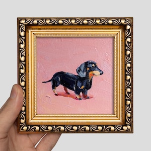Op de afbeelding: Een ingelijst olieverfschilderij van een teckel. De hond is zwart en bruin, tegen een roze achtergrond. Het schilderij zit in een gouden, sierlijke lijst, vastgehouden door een hand.