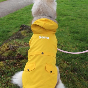 May include: A yellow dog raincoat with a hood and two pockets. The raincoat has the name "Sora" embroidered on the back.