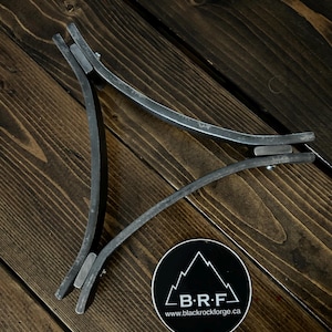 May include: A metal tripod stand with three curved legs. The stand is made of black metal and has a simple design. The stand is on a wooden surface. The stand is made by Black Rock Forge.