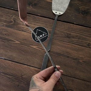 Peut inclure: Deux spatules en métal forgées à la main avec de longs manches fins. Une spatule présente une lame couleur cuivre, tandis que l'autre possède une lame en acier texturé sombre fixée par deux rivets. Un autocollant circulaire avec le texte 3 R F blackrockforge.ca est visible.