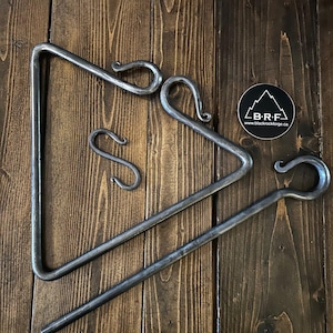 May include: A set of three black metal fireplace tools, including a triangle stand, a poker, and two hooks. The tools are arranged on a wooden surface. The tools are made by Blackrock Forge, as indicated by the sticker in the image.