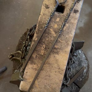 Könnte beinhalten: Zwei handgeschmiedete Metall-Schürhaken mit gedrehten Schäften und kreisförmigen Griffschlaufen am oberen Ende. Jeder Schürhaken hat einen schlanken, dunkelgrauen Eisenkörper mit einem kleinen Haken am unteren Ende, der auf einem Amboss liegt.