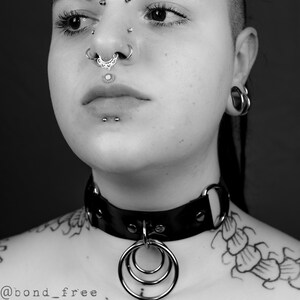 K&ouml;nnte beinhalten: Eine Person mit kurzen schwarzen Haaren und mehreren Gesichtspiercings tr&auml;gt eine schwarze Lederkette mit einem silbernen Ring. Die Person hat ein Tattoo auf der Schulter.