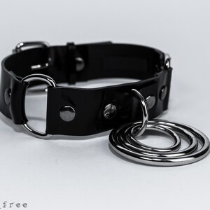 K&ouml;nnte beinhalten: Ein schwarzes Lederhalsband mit silbernen Metallbeschl&auml;gen, darunter ein gro&szlig;er Ring mit drei kleineren Ringen. Das Halsband hat einen Schnallenverschluss und mehrere silberne Nieten.
