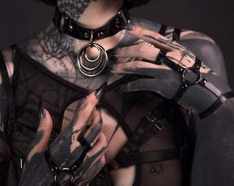 Puede incluir: Una persona con tatuajes de tinta negra lleva un collar de cuero negro con un colgante de anillo plateado y puños de cuero negro con anillos plateados.