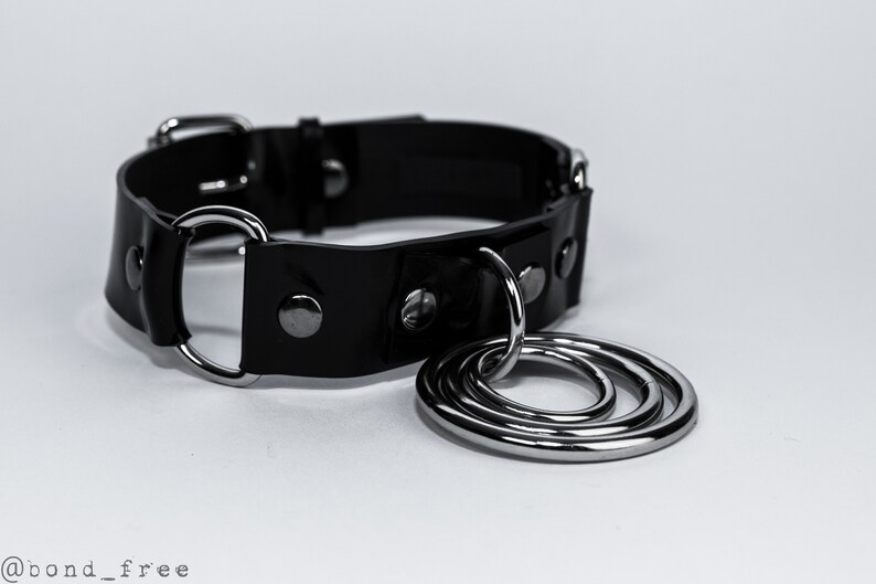 K&ouml;nnte beinhalten: Ein schwarzes Lederhalsband mit silbernen Metallbeschl&auml;gen, darunter ein gro&szlig;er Ring mit drei kleineren Ringen. Das Halsband hat einen Schnallenverschluss und mehrere silberne Nieten.