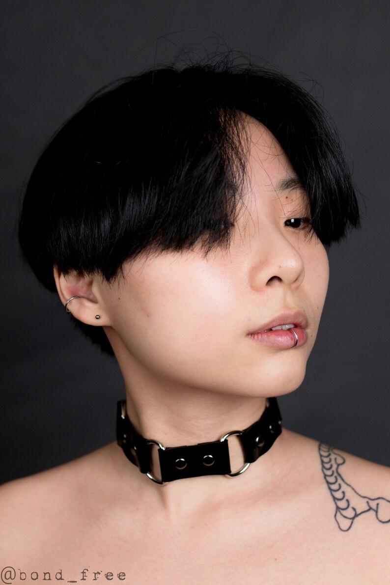May include: A black leather choker necklace with silver-colored metal rings and studs. The choker is worn around the neck. The image also shows a person with black hair, a lip piercing, and a tattoo on the shoulder.