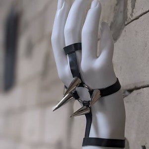 Puede incluir: Un brazalete de cuero negro con pinchos plateados y un anillo. El brazalete es ajustable y tiene una est&eacute;tica punk rock.