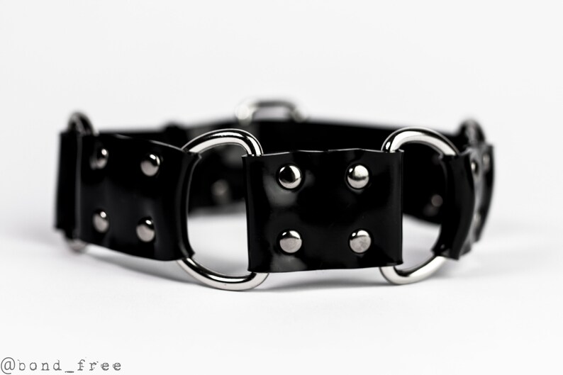 May include: A black, glossy collar with silver-toned metal rings and rivets. The collar is made of a shiny material and features a simple, yet bold design. The image is set against a white background.