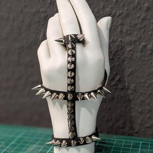 K&ouml;nnte beinhalten: Ein schwarzes Lederarmband mit silbernen Spikes. Das Armband ist so konzipiert, dass es am Handgelenk und an der Hand getragen werden kann, mit einem Band, das um das Handgelenk geht, und einem zweiten Band, das um die Hand geht. Das Armband ist mit silbernen Spikes verziert, die in einem Muster angeordnet sind.