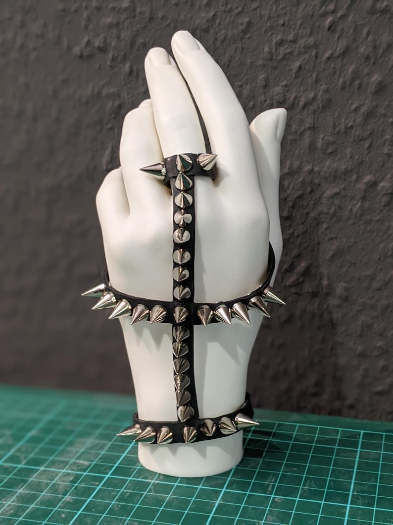 K&ouml;nnte beinhalten: Ein schwarzes Lederarmband mit silbernen Spikes. Das Armband ist so konzipiert, dass es am Handgelenk und an der Hand getragen werden kann, mit einem Band, das um das Handgelenk geht, und einem zweiten Band, das um die Hand geht. Das Armband ist mit silbernen Spikes verziert, die in einem Muster angeordnet sind.