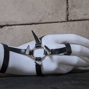 Puede incluir: Un brazalete de cuero negro con p&uacute;as plateadas y un anillo. El brazalete se lleva en una mano de maniqu&iacute; blanca.