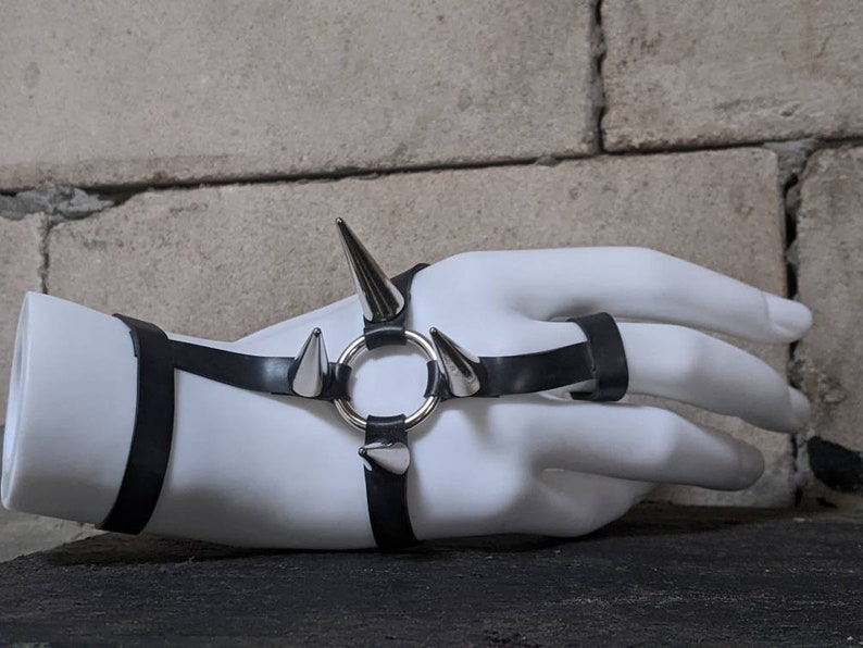Puede incluir: Un brazalete de cuero negro con p&uacute;as plateadas y un anillo. El brazalete se lleva en una mano de maniqu&iacute; blanca.