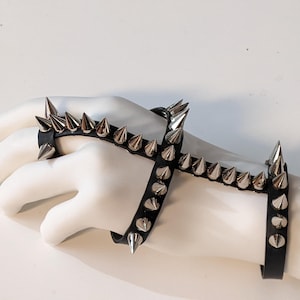 K&ouml;nnte beinhalten: Ein schwarzes Lederarmband mit silbernen pyramidenf&ouml;rmigen Nieten. Das Armband wird an einer Mannequinhand getragen und umschlie&szlig;t Handgelenk und Finger.