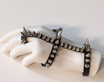 Puede incluir: Un brazalete de cuero negro con tachuelas piramidales plateadas. El brazalete se lleva en una mano de maniquí y se envuelve alrededor de la muñeca y los dedos.