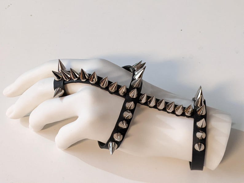 K&ouml;nnte beinhalten: Ein schwarzes Lederarmband mit silbernen pyramidenf&ouml;rmigen Nieten. Das Armband wird an einer Mannequinhand getragen und umschlie&szlig;t Handgelenk und Finger.