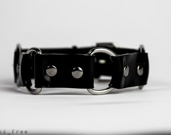 Puede incluir: Un collar negro con anillos y remaches de metal plateado. El collar está hecho de un material brillante, similar al charol. El diseño incluye múltiples anillos circulares y un cierre de hebilla. El producto se muestra sobre un fondo blanco.