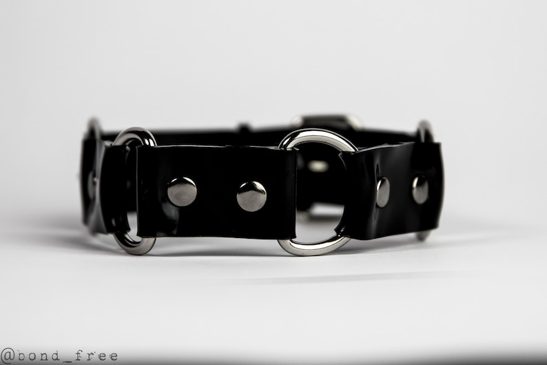 May include: A black collar with silver-colored metal rings and rivets. The collar is made of a shiny, patent-like material. The design includes multiple circular rings and a buckle closure. The product is displayed against a white background.