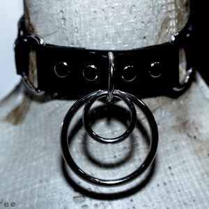 Puede incluir: Collar de gargantilla de cuero negro con herrajes de metal plateado y dos anillos entrelazados. El collar se exhibe en una cabeza de maniqu&iacute;. El dise&ntilde;o es simple y elegante.