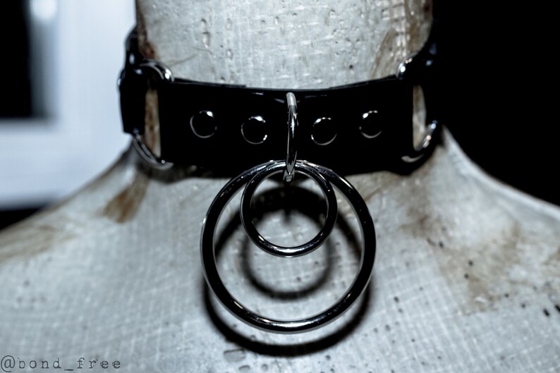Puede incluir: Collar de gargantilla de cuero negro con herrajes de metal plateado y dos anillos entrelazados. El collar se exhibe en una cabeza de maniqu&iacute;. El dise&ntilde;o es simple y elegante.
