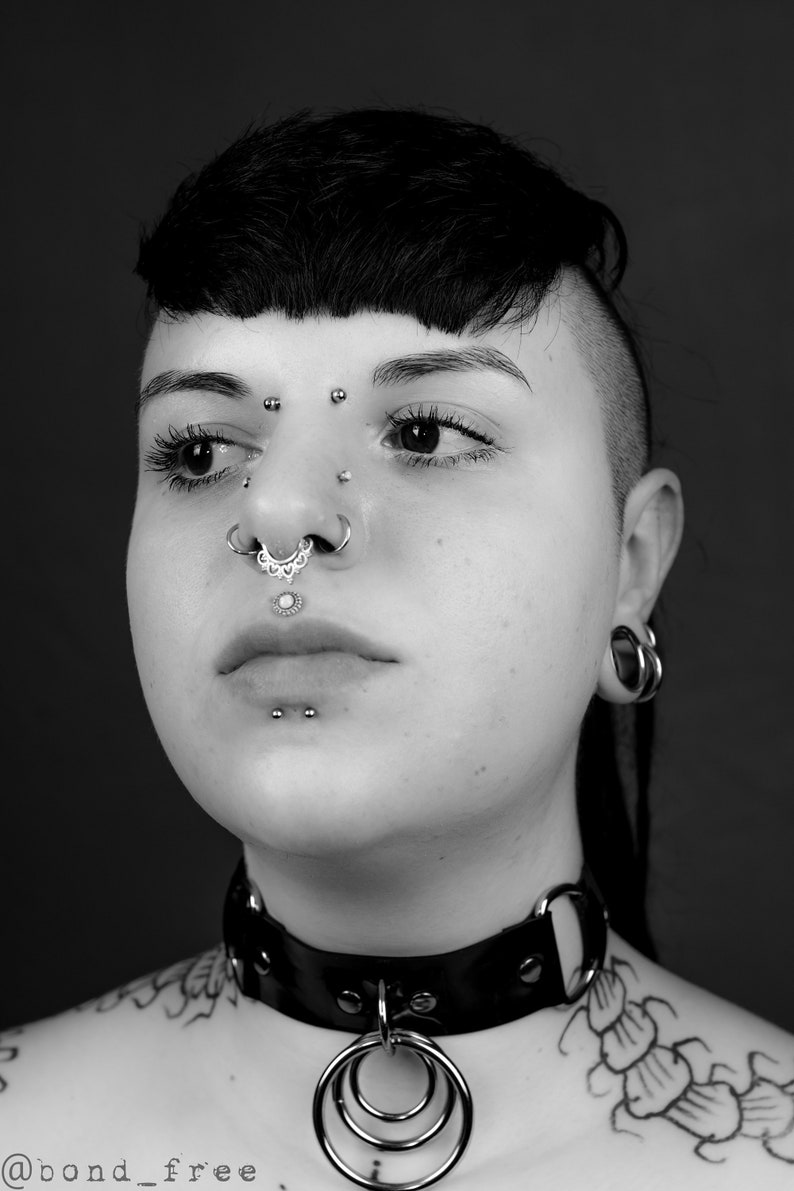 K&ouml;nnte beinhalten: Eine Person mit kurzen schwarzen Haaren und mehreren Gesichtspiercings tr&auml;gt eine schwarze Lederkette mit einem silbernen Ring. Die Person hat ein Tattoo auf der Schulter.