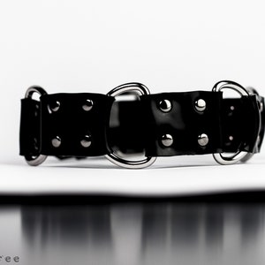 May include: A black, glossy collar with silver-toned metal rings and rivets. The collar is made of a flexible material and is designed with a series of connected segments. The image is set against a white background.
