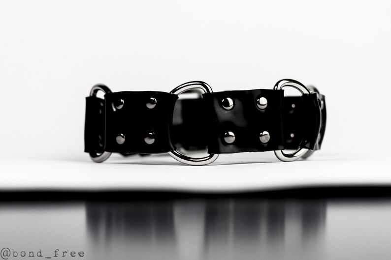 May include: A black, glossy collar with silver-toned metal rings and rivets. The collar is made of a flexible material and is designed with a series of connected segments. The image is set against a white background.
