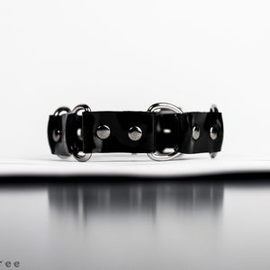 May include: A black leather collar with silver-toned metal hardware. The collar features a series of circular rings and small, round studs. The design is simple and sleek, suitable for various styles.