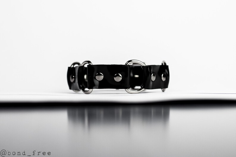 May include: A black leather collar with silver-toned metal hardware. The collar features a series of circular rings and small, round studs. The design is simple and sleek, suitable for various styles.
