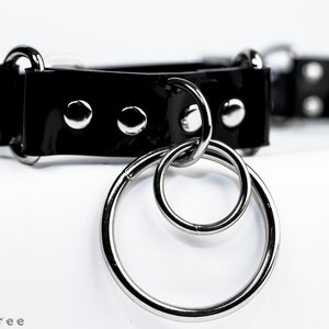 Puede incluir: Un collar tipo gargantilla negro brillante con herrajes met&aacute;licos plateados. El collar presenta tres tachuelas plateadas y un par de anillos entrelazados. El fondo es blanco.