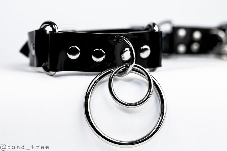 Puede incluir: Un collar tipo gargantilla negro brillante con herrajes met&aacute;licos plateados. El collar presenta tres tachuelas plateadas y un par de anillos entrelazados. El fondo es blanco.