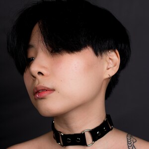 May include: A black leather choker necklace with silver-colored metal rings and rivets. The choker is worn around the neck. The person in the image has short black hair and a lip piercing. The background is a solid dark gray.