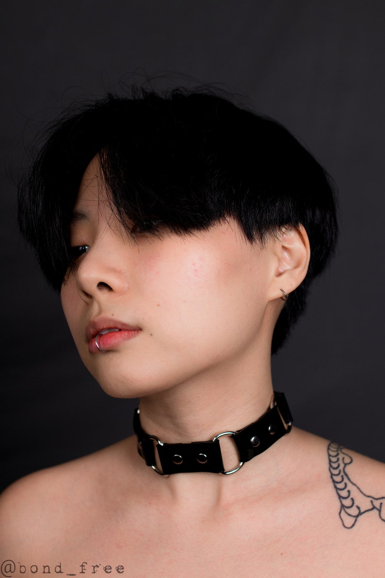 May include: A black leather choker necklace with silver-colored metal rings and rivets. The choker is worn around the neck. The person in the image has short black hair and a lip piercing. The background is a solid dark gray.