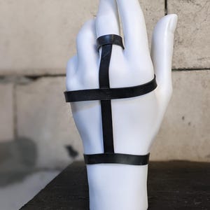 May include: A black leather ring with a cross design. The ring is worn on a mannequin hand.