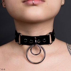 Puede incluir: Collar gargantilla de cuero negro con herrajes de metal plateado y dos anillos entrelazados. La gargantilla tiene forma rectangular con esquinas redondeadas y se asegura con una hebilla. El dise&ntilde;o es minimalista y atrevido.