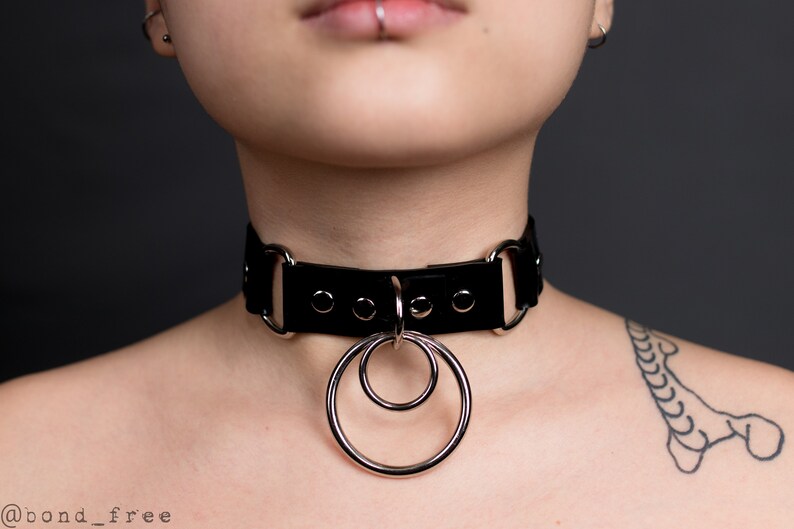 Puede incluir: Collar gargantilla de cuero negro con herrajes de metal plateado y dos anillos entrelazados. La gargantilla tiene forma rectangular con esquinas redondeadas y se asegura con una hebilla. El dise&ntilde;o es minimalista y atrevido.