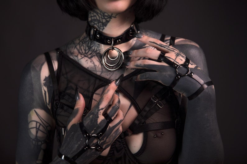 K&ouml;nnte beinhalten: Eine Person mit schwarzen Tattowierungen tr&auml;gt eine schwarze Lederkette mit einem silbernen Ringanh&auml;nger und schwarze Lederarmb&auml;nder mit silbernen Ringen.