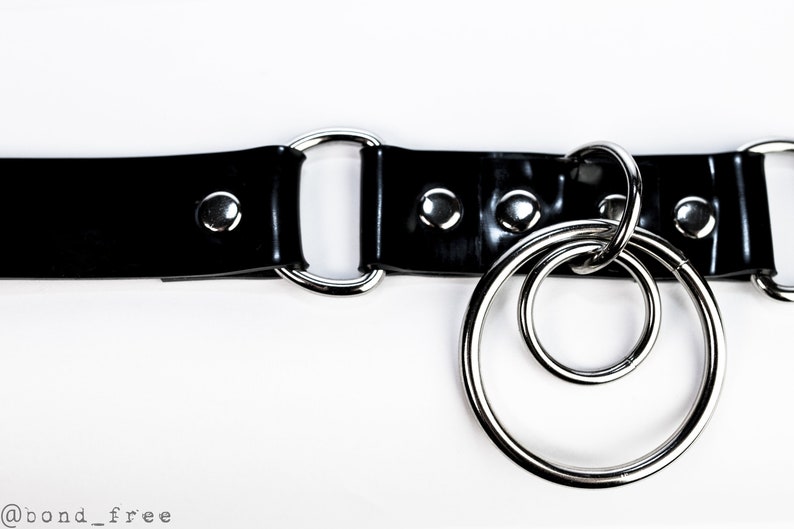 Puede incluir: Un cintur&oacute;n negro brillante con herrajes met&aacute;licos plateados. El cintur&oacute;n presenta una serie de remaches y dos grandes anillos circulares conectados. El fondo es blanco.