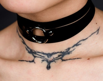 Puede incluir: Collar gargantilla negro con un diseño de doble banda y un anillo plateado en el centro. El collar se lleva alrededor del cuello. La imagen también muestra un tatuaje en el cuello y el pecho.