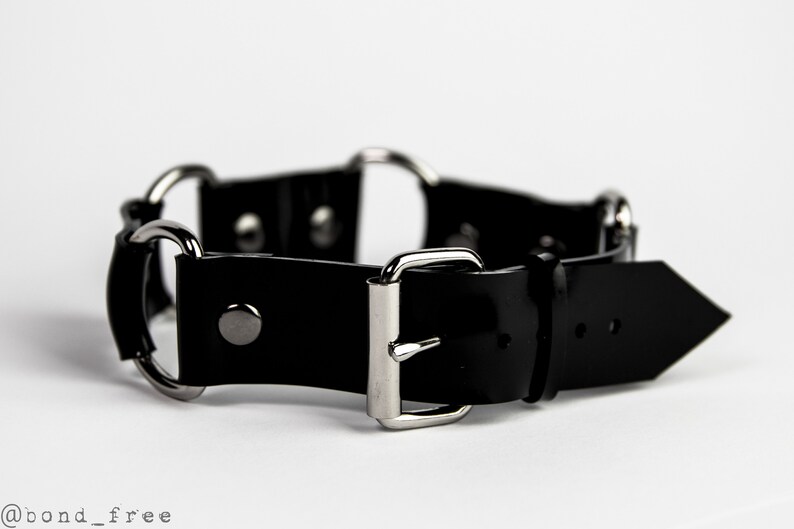 May include: A black leather collar with silver-toned metal hardware. The collar features a buckle closure and several metal rings. The design is simple and sleek, with a focus on the contrast between the black leather and the silver metal.