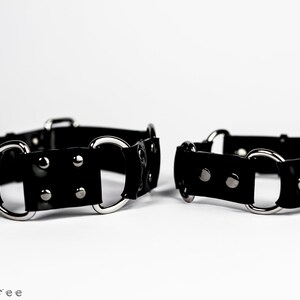 May include: Two black leather wrist cuffs with silver-colored metal rings and rivets. The cuffs are displayed on a white background. The cuffs are a fashion accessory.