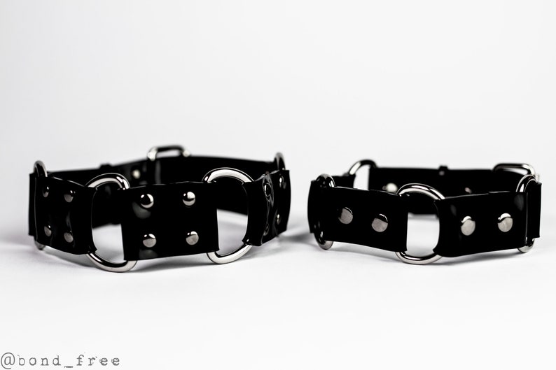 May include: Two black leather wrist cuffs with silver-colored metal rings and rivets. The cuffs are displayed on a white background. The cuffs are a fashion accessory.