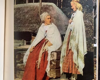 May include: Two people in traditional clothing, likely from a historical or cultural context. One person is seated, wearing a red plaid skirt, white shawl, and head covering. The other person is standing, also in a plaid skirt and shawl, with a decorative headpiece. The background includes a rustic building and trees.