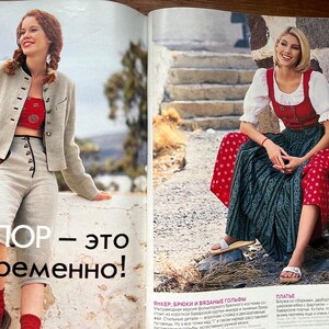 May include: Two women in traditional folk attire. One is wearing a light grey jacket, red top, and light grey trousers. The other is in a red vest over a white blouse and a long green skirt with red polka dots.