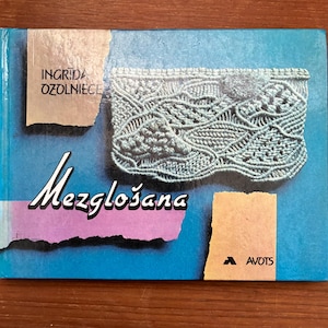 May include: A book cover with the title "Mezglošana" in a cursive font, featuring a detailed macrame design. The cover has a blue background with the author's name, Ingrid Ozolniece, and the AVOTS logo.