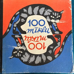Könnte beinhalten: Ein Vintage-Buchcover mit zwei stilisierten schwarzen und grauen Katzen mit weißen Schnurrhaaren, vor einem roten und blauen Hintergrund. Der Buchtitel "100 mīklu" ist in blauer und roter Schrift geschrieben.
