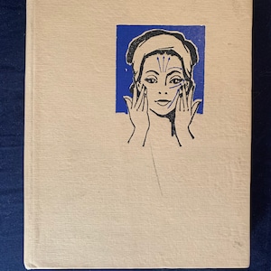 May include: A vintage book with a cream-coloured cover. A blue square on the cover features a black and white illustration of a person's face with hands touching the face. The person is wearing a head wrap, and lines indicate facial massage techniques.