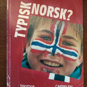 Puede incluir: Portada de libro roja con el título "TYPISK NORSK?" en letras blancas. La portada presenta a una persona sonriente con la bandera noruega pintada en la cara. El libro se titula "Tekstbok" y es publicado por Cappelen.