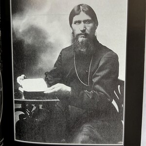 May include: A black and white photograph of Grigori Rasputin, seated and holding a book. He has a long beard and is wearing a dark-colored garment. The image is framed within a black border, with text at the bottom.