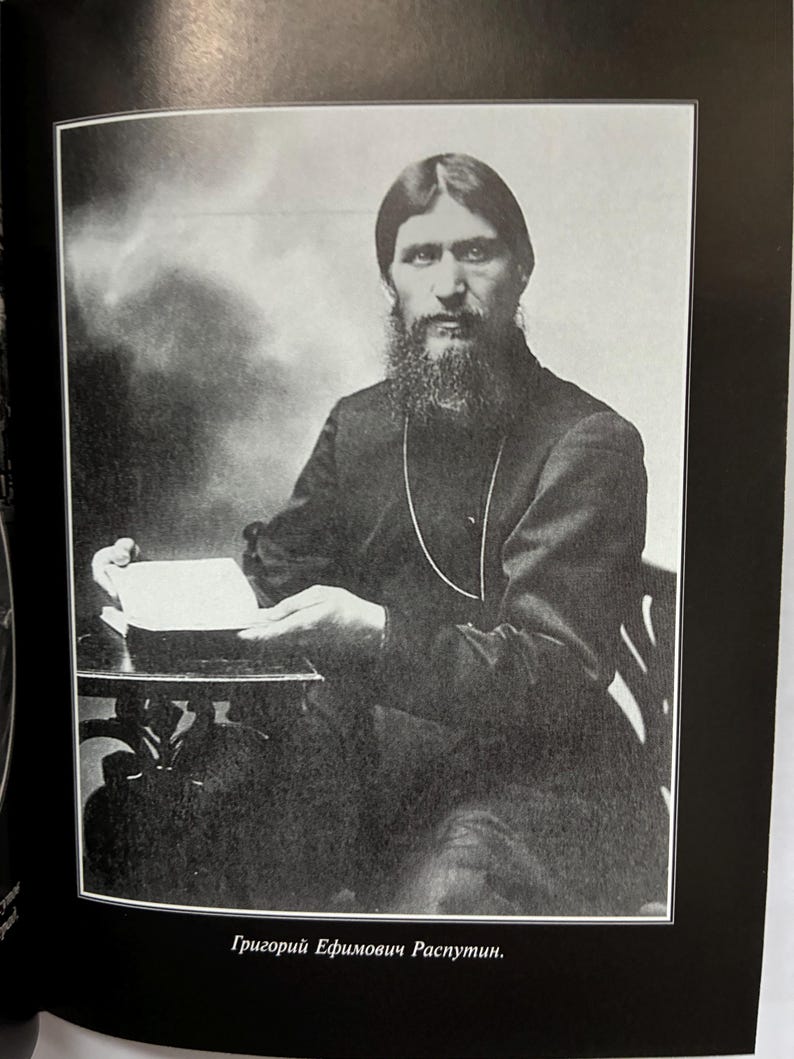 May include: A black and white photograph of Grigori Rasputin, seated and holding a book. He has a long beard and is wearing a dark-colored garment. The image is framed within a black border, with text at the bottom.